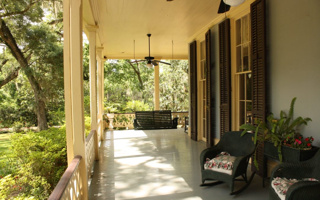 A porch with comfortable seating, including a swing and chairs, surrounded by lush greenery and trees, creating a welcoming outdoor space.